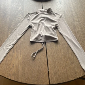 Beige ribbad croppad topp med snörning  - Trendig beige croppad topp med lång ärm och hög krage. Toppen har snygg snörning i ryggen och är tillverkad i ett ribbat, stretchigt material som sitter snyggt på kroppen.
