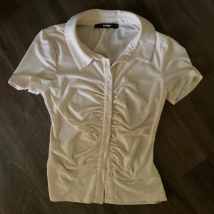 Beige kortärmad ”skjorta” med rynk från Bik Bok - Snygg beige skjorta från Bik Bok med korta ärmar och knappar framtill. Skjortan har rynkade detaljer längs knappraden som ger en cool och trendig look. Perfekt till jeans eller kjol för en fräsch stil. Det finns två super små fläckar som säkert kan tvättas bort🌸🤍