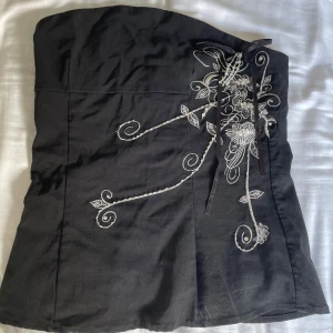 Svart tubtopp - Säljer en svart tubtopp från ICHI med vackert vitt broderi och snörning framtill. Toppen har blommiga detaljer och är axelbandslös, perfekt för att skapa en snygg och unik look. Använd ettt fåtal gånger. Jag är en S och kan använda den även om det är en M. Säljs pågrund av att den har tyvärr blivit för liten!❤️