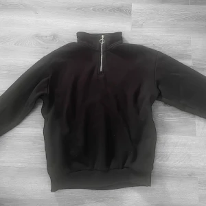 Svart tröja med dragkedja från Bershka - Svart tröja från Bershka i storlek S. Tröjan har hög krage och en dragkedja framtill med rund metallring som detalj. Perfekt för en stilren och enkel look. Skriv vid funderingar