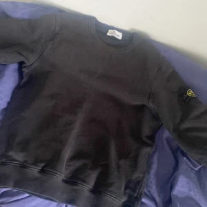 Svart sweatshirt från Stone Island Junior, passar 158cm - Säljer en svart sweatshirt från Stone Island Junior med klassisk rund halsringning och logotyp-patch på vänster ärm. Tröjan har en enkel och stilren design, 