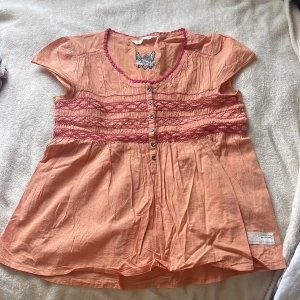 Persikofärgad blus med broderi från Odd Molly - Säljer en persikofärgad blus från Odd Molly med korta ärmar, dekorativa röda broderier och knappar framtill. Blusen har en söt fjärilsbrodyr på ryggen och är tillverkad i mjuk bomull. Perfekt för vår och sommar!