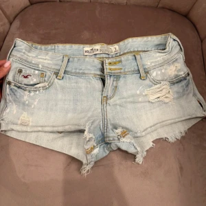 Ljusblå jeansshorts från Hollister - Säljer ett par ljusblå jeansshorts från Hollister med slitna detaljer, fransig kant och klassiska fickor både fram och bak. Shortsen har låg midja och gult broderi på bakfickorna. Perfekta för varma dagar! 🩵🩵