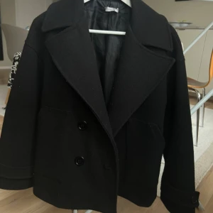 Svart kappa  - Stilren svart kappa i ull med stora slag och dubbelknäppning framtill. Jackan har långa ärmar och två stora fickor framtill. Perfekt för kyligare dagar och ger en klassisk look. Den är lite nopprig 