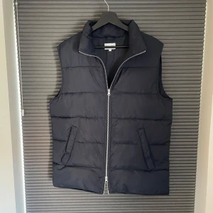 Marinblå dunväst  - Marinblå dunväst från East West med quiltad design och tvåvägsdragkedja framtill. Västen har två snedställda fickor på utsidan och en innerficka med dragkedja. Använd fåtal gånger mycket bra skick.