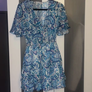 mönstrad kort klänning från Zara - Säljer en superfin kort klänning från Zara med blått och lila mönster. Klänningen har v-ringning, volangdetaljer och korta ärmar. Den är gjord i ett lätt, luftigt material och har resår i midjan för en snygg passform. Perfekt för sommarens alla tillfällen!