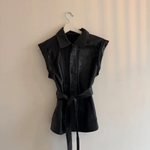 Svart väst i skinnimitation med bälte - snygg väst i svart läderimitation. Den stängs med dragkedja framtill och har ett knytband i midjan som ger en fin och markerad siluett. OBS: Vet inte storlek men skulle uppskatta XS/S.  