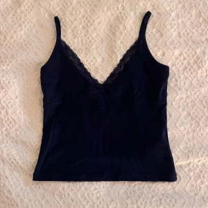 Mörkblå linnetopp med spetskant - Supersnygg mörkblå linnetopp med tunna axelband och v-ringning dekorerad med spets. Toppen har en enkel och stilren design som passar perfekt till jeans eller kjol. Använd fåtals gånger 