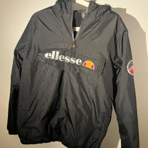 Svart vindjacka från Ellesse - Svart vindjacka från Ellesse med stor logga framtill och patch på ena ärmen. Jackan har huva, halv dragkedja och en stor ficka på magen. Perfekt för dig som vill ha en sportig och clean stil. Materialet är lätt och känns smidigt.