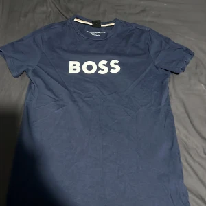Marinblå BOSS t-shirt XS - Snygg marinblå t-shirt från BOSS i bomull med klassisk passform. Stor vit BOSS-logga på bröstet ger en clean och modern look. Perfekt för dig som gillar stilrena plagg med tydlig branding. Rund halsringning och korta ärmar.