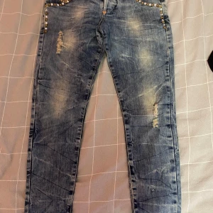 Blå slitna jeans från Justor med nitar - Snygga blå jeans från Justor, Made in Italy, med slitna detaljer och coola nitar vid fickorna. Jeansen har en tvättad look och raka ben. Perfekta för dig som gillar en edgy stil med extra detaljer.