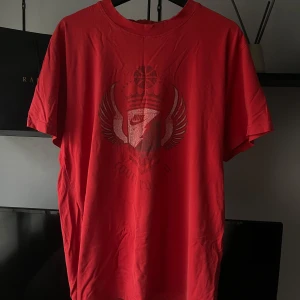Röd Nike t-shirt  - Snygg röd t-shirt från Nike med coolt grafiskt tryck på bröstet som inkluderar vingar, krona och basketboll. Klassisk rund hals och korta ärmar. Tillverkad i mjuk bomull som är skön mot huden. Perfekt för dig som gillar sportig streetstyle.