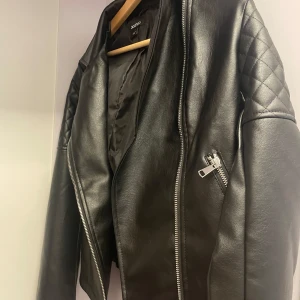 Svart skinnjacka från Boohoo - Snygg svart skinnjacka från Boohoo med quiltade detaljer på axlarna och silvriga dragkedjor. Jackan har en klassisk bikerstil och är perfekt för dig som vill ha en edgy look. Fodrad insida och två fickor med dragkedja framtill.