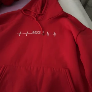 Röd hoodie med 2023-tryck - Säljer en röd hoodie med vit tryck där det står 2023 och ett coolt pulsmönster över bröstet. Tröjan har huva med snörning och en stor magficka framtill. Perfekt för dig som gillar streetwear och vill sticka ut med färg.