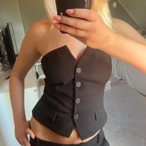 Svart tub-topp med knappar - Snygg svart tub-topp  med knappar framtill och markerad midja. Toppen är axelbandslös och har en cool, skräddad look med fickdetaljer och smockad rygg för extra passform. Perfekt till kostymbyxor eller jeans för en trendig vibe.