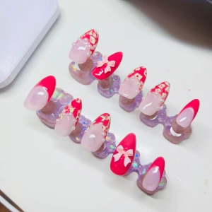 Rosa och röda press-on naglar med rosett - Säljer ett set med press-on naglar i mandelform med rosa och röd design. Några naglar har handmålade blommor och pärlor, andra har en söt 3D-rosett. Perfekt för dig som vill ha en unik och färgglad look på naglarna.