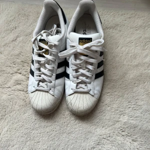 Adidas Superstar vita sneakers - Klassiska Adidas Superstar sneakers i vitt skinn med svarta ränder och gulddetaljer på plösen. Skorna har snörning, rund tå och den ikoniska gummitåhättan. Perfekta för en streetstyle-look och funkar året runt.