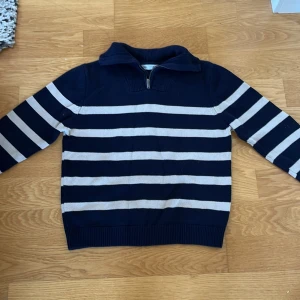 Marinblå randig stickad tröja Zara - Snygg stickad tröja från Zara i marinblått med vita horisontella ränder. Tröjan har en hög krage med half zip-dragkedja och långa ärmar. Perfekt för dig som gillar klassisk stil med en modern twist.