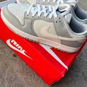 Nike Dunk Low  - Hej! Säljer ett par Nike Dunk Low sneakers i en clean ljusgrå och vit färg som jag fick av min farfar i sen födelsedagspresent. Skorna har en rund tå, vit snörning och platt sula.  Ovandelen är tillverkad i slät och texturerad skinn med klassisk Nike Swoosh på sidan.  Skorna är bara provade och jag gillade inte dom, därför säljer jag dom!  Vill du veta något mer eller vill ha fler bilder så är det bara att skriva! Priset är såklart inte hugget i sten!
