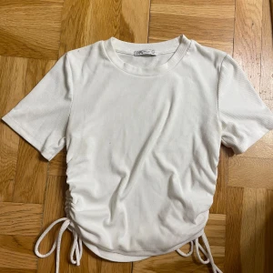 Vit ribbad topp med snörning från Zara - Supersnygg vit ribbad topp från Zara med korta ärmar och rund hals. Toppen har trendig snörning i sidorna som gör att du kan justera passformen och skapa en cool, rynkad effekt. OBS! Liten i storlek (passar M). Lite nopprig i materialet men inget man lägger märke till