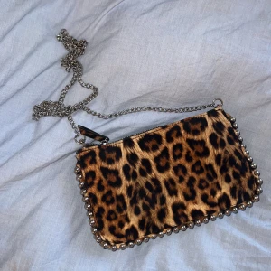 Leopardmönstrad kuvertväska med kedja - Snygg leopardmönstrad kuvertväska med silverfärgad kedja och dragkedja. Perfekt för att ge din outfit en vild touch. Väskan har dekorativa nitar längs kanterna som ger en extra edge. Kan skicka fler bilder på den. I Nyskick