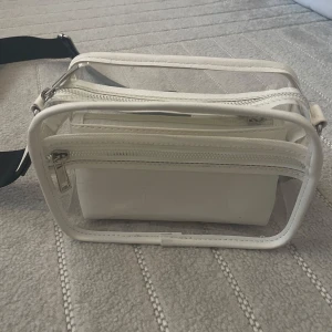 Transparent axelväska med vit mindre väska. - Säljer en transparent axelväska med en vit mindre väska inuti med egen dragskedja som går att ta bort. Väskan har en svart justerbar axelrem och dragkedja upptill.