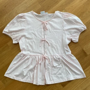 Ljusrosa blus med knyt från Gina Tricot - Supersöt ljusrosa blus från Gina Tricot med kort puffärm och tre dekorativa knyt framtill. Modellen har en lätt utställd nederdel och rund halsringning. Använd en gång. Du står för frakten, vid mer info skriv 