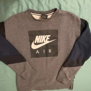Grå Nike Air sweatshirt med dragkedja - Säljer en grå Nike Air sweatshirt i storlek S med svart och marinblåa paneler på ärmarna. Stor Nike Air-logga på bröstet och en cool dragkedja längs sidan. Mjuk insida i fleece och klassisk rund hals. Perfekt för dig som gillar sportig streetstyle.