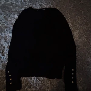 Svart stickad tröja från Zara - Svart stickad tröja från Zara i storlek S. Tröjan har långärmade ärmar med snygga silvriga knappar vid ärmsluten som ger en cool detalj. Perfekt att matcha med jeans eller kjol för en stilren look.