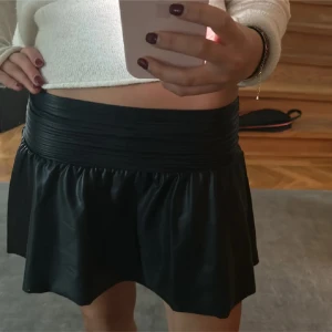 Svart kort kjol - Säljer en svart kort kjol med bred, rynkad midja och lätt utsvängd form. Kjolen är gjord i skinnimitation. Perfekt att matcha med en stickad tröja eller boots för en trendig look.