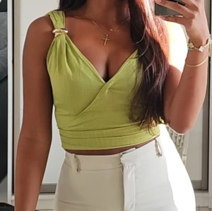 Ljusgrön omlott croppad linnetopp - Snygg ljusgrön croppad linnetopp med omlott-skärning och breda axelband dekorerade med guldfärgade ringar. Toppen har djup v-ringning och sitter tight över kroppen, perfekt till höga byxor eller kjol.
