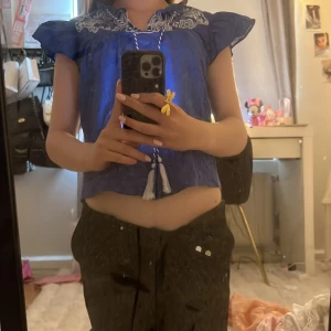Blå blus med broderi och volangärm - Supersöt blå blus med vita broderier vid halsen och volangärmar. Blusen har knytband med tofsar framtill och är lätt och luftig i materialet. Perfekt för dig som gillar unika detaljer och en chill vibe.