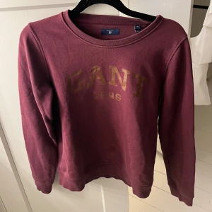 Vinröd sweatshirt från GANT XS - Vinröd sweatshirt från GANT i storlek XS med guldig logga och årtal tryckt framtill. Klassisk rund halsringning och långa ärma, väldigt skön. Har en flott fläck i fram som inte försvunnit i tvätten, syns på första och sista bilden.
