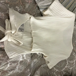 Vit ribbad omlottopp med knyt - Superfin vit ribbad omlottopp från H&M med korta ärmar och knyt i sidan. Toppen har en mjuk och stretchig känsla och är perfekt för att skapa en chill men ändå snygg look. Passar till både jeans och kjol för en trendig vibe. Liten fläck på framsidan men syns knappt 