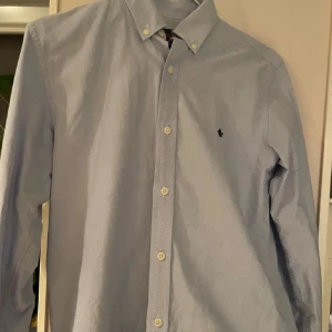 Ljusblå skjorta från morris - Klassisk ljusblå skjorta från morris med button down-krage och broderad logga på bröstet. Skjortan har långa ärmar och vita knappar framtill. Perfekt för en clean och stilren look.