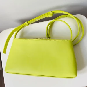 Limegul axelväska i syntet - Säljer en stilren axelväska i limegul färg med justerbart axelband och dragkedja upptill. Väskan har en minimalistisk design och är tillverkad i slätt syntetmaterial. Perfekt för dig som vill sticka ut med en färgklick i din outfit.