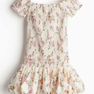 Vit blommig offshoulder kortklänning - Supersöt vit kortklänning med blommigt mönster i rosa och gult. Offshoulder med korta ärmar och volang nedtill som ger en romantisk vibe. Smockad överdel och luftig kjol, perfekt för varma dagar. Passar dig som gillar feminin och trendig stil.