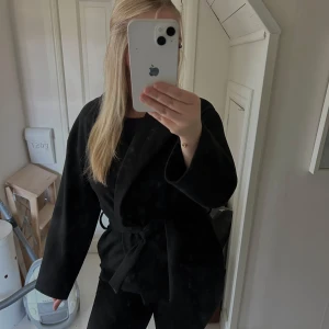 Svart omlott kappa med bälte - Säljer en svart kappa med omlottmodell och bälte i midjan. Jackan har en loose passform och långa ärmar, perfekt för dig som gillar en stilren och enkel look. Materialet känns mjukt och ger en snygg siluett. Passar till många olika outfits. I bra skick därav använd fåtal gånger.