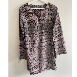 Blommig kortklänning från Rut&Circle - Säljer en svart och ljusrosa kortklänning från Rut&Circle med blommigt mönster och snörning vid halsen. Klänningen har långärmade ärmar och är tillverkad i ett mjukt, lätt material som ger en skön känsla. Perfekt för dig som gillar boho-vibe och detaljer.