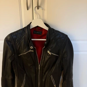 Svart skinnjacka Rock N Blue, stl 34 - Snygg svart skinnjacka från Rock N Blue med coola quiltade detaljer på axlarna och flera dragkedjor framtill. Röd insida som ger extra edge. Jackan har en figurnära passform och klassisk bikerstil. Perfekt för dig som vill ha en stilren och tuff look.