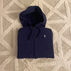 Ralph Lauren zip hoodie – Marinblå | Storlek: L (passar mer som M) - Klassisk och stilren marinblå zip hoodie från Ralph Lauren med vit broderad logga på bröstet. Mjuk och bekväm bomullsblandning, perfekt för vardagsbruk eller som ett extra lager. Huvan har justerbara snören med metalländar och dragkedja framtill.  ✔ Färg: Marinblå ✔ Passform: Normal till lite slim (mer som M än L) ✔ Skick: Mycket bra, sparsamt använd