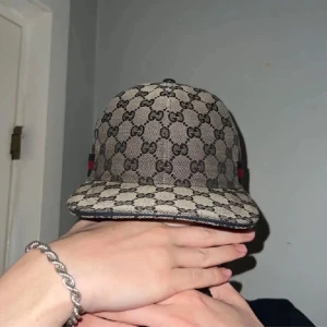 Gucci keps med GG-mönster och läderdetalj - Snygg Gucci keps i beige med klassiskt GG-monogram över hela kepsen. Har justerbar rem i svart skinn baktill och ikoniska röd-blå ränder på sidan. Tillverkad i Italien och perfekt för dig som vill sticka ut med lyxig streetstyle.