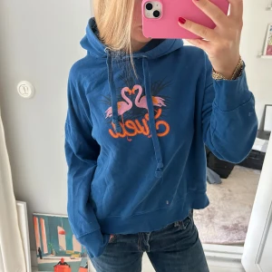 Blå hoodie med flamingotryck - Säljer en blå hoodie från Chiquelle med coolt flamingotryck i rosa och orange på bröstet. Tröjan har huva med snörning och långa ärmar, perfekt för chill dagar. 
