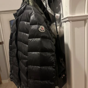 Svart Moncler Acourus - Säljer en svart Moncler Acourus med klassisk quiltad design och diskret logga på ärmen. Jackan har dragkedja framtill, hög krage och två fickor med dragkedja. Perfekt för dig som vill ha en lätt men varm jacka med stilren look. Storleken är 3 (passar M/L)