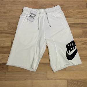 Nike shorts vita - Snygga vita shorts från Nike med stor svart logga på vänster ben. Modellen har elastisk midja med snörning, två sidofickor och en bakficka. Mjukt bomullsmaterial som är skönt mot huden och passar perfekt till chill eller sport.