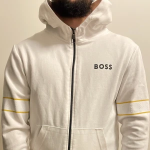 Vit zip-hoodie från BOSS med gula detaljer - Vit hoodie från BOSS med dragkedja framtill och huva. Ärmarna har gula ränder och präglad BOSS-logga. Mjuk bomullskänsla och fickor framtill. Perfekt för en clean och sportig look.