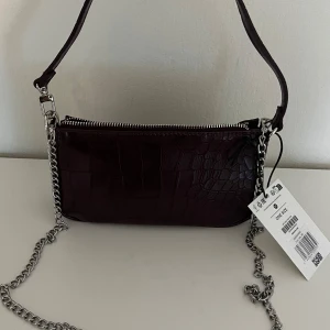 Mörklila axelväska med kedja - Snygg mörklila axelväska med croco-mönster och silverfärgad kedja som axelrem. Väskan har dragkedja upptill och en stilren, rektangulär form. Perfekt för att lyfta din outfit med en cool detalj. Materialet är skinnimitation och väskan har en modern vibe. OBS väskan är från ASOS och aldrig använd, någon basic väska och aldrig använd))