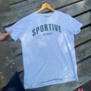 Grå t-shirt - Ljusgrå t-shirt med trycket 'SPORTIVE UNIVERSITY' i grönt.