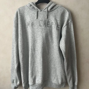 Grå hoodie  - Snygg ljusgrå hoodie från Gina Tricot med texten 'MA CHÉRIE' i glitter på bröstet. Tröjan har huva, långa ärmar och ribbade muddar. Perfekt för en chill och trendig look. Endast använd 1 gång.