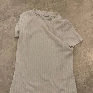 Ribbad vit t-shirt från Lindex - Vit ribbad t-shirt från Lindex med korta ärmar och rund hals. T-shirten har en clean och enkel look med tunna vertikala ränder i materialet. Perfekt för dig som gillar basic och stilrena plagg.